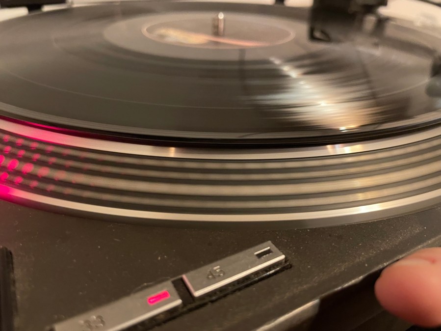 A record spinning on a Technics SL-1210 record player. The light is reflecting off the grooves of the black vinyl and in the foreground the speed controller is set to 33 1/3 RPM